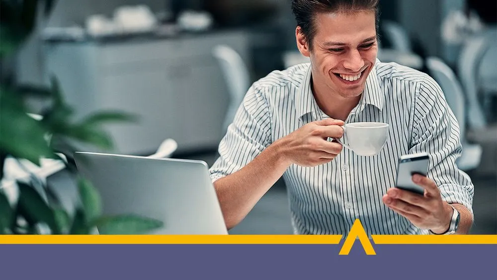 uśmiechnięty mężczyzna w koszuli siedzący przy biurku, trzymający filiżankę kawy i korzystający z telefonu