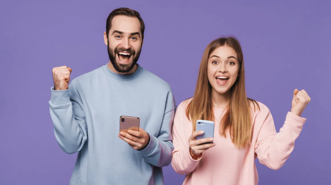 Uśmiechnięty mężczyzna i kobieta w pastelowych bluzach, trzymający smartfony i unoszący zaciśnięte pięści w geście radości na tle fioletowej ściany.